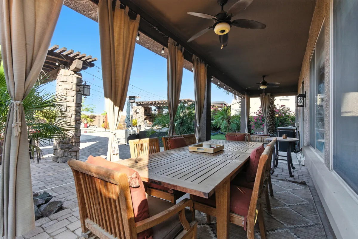 A relaxed outdoor dining area with a sturdy wooden table, flowing curtains, an outdoor ceiling fan, and stone pillars surrounded by lush greenery. The scene is set for al fresco meals and social evenings under the stars.