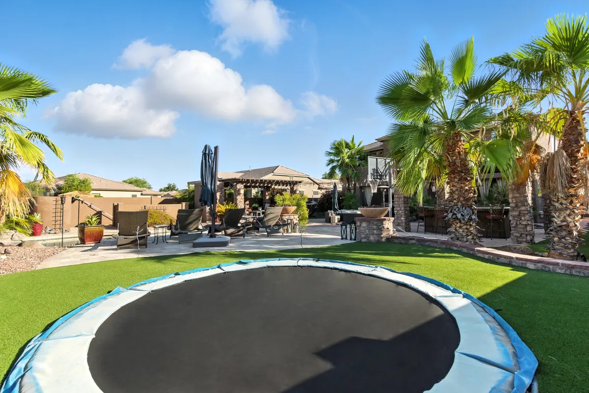 A wide view of the backyard featuring a sparkling swimming pool, a built-in in-ground trampoline, outdoor seating, and tall palm trees. The scene captures spaces for both relaxation and play, with the pool inviting for swims and the trampoline promising fun for kids and adults alike.