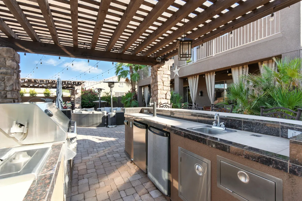 A lively and well-appointed outdoor kitchen under a pergola, with ample seating and lush landscaping all around. The space is set up for entertaining and al fresco dining, with string lights adding a festive touch.