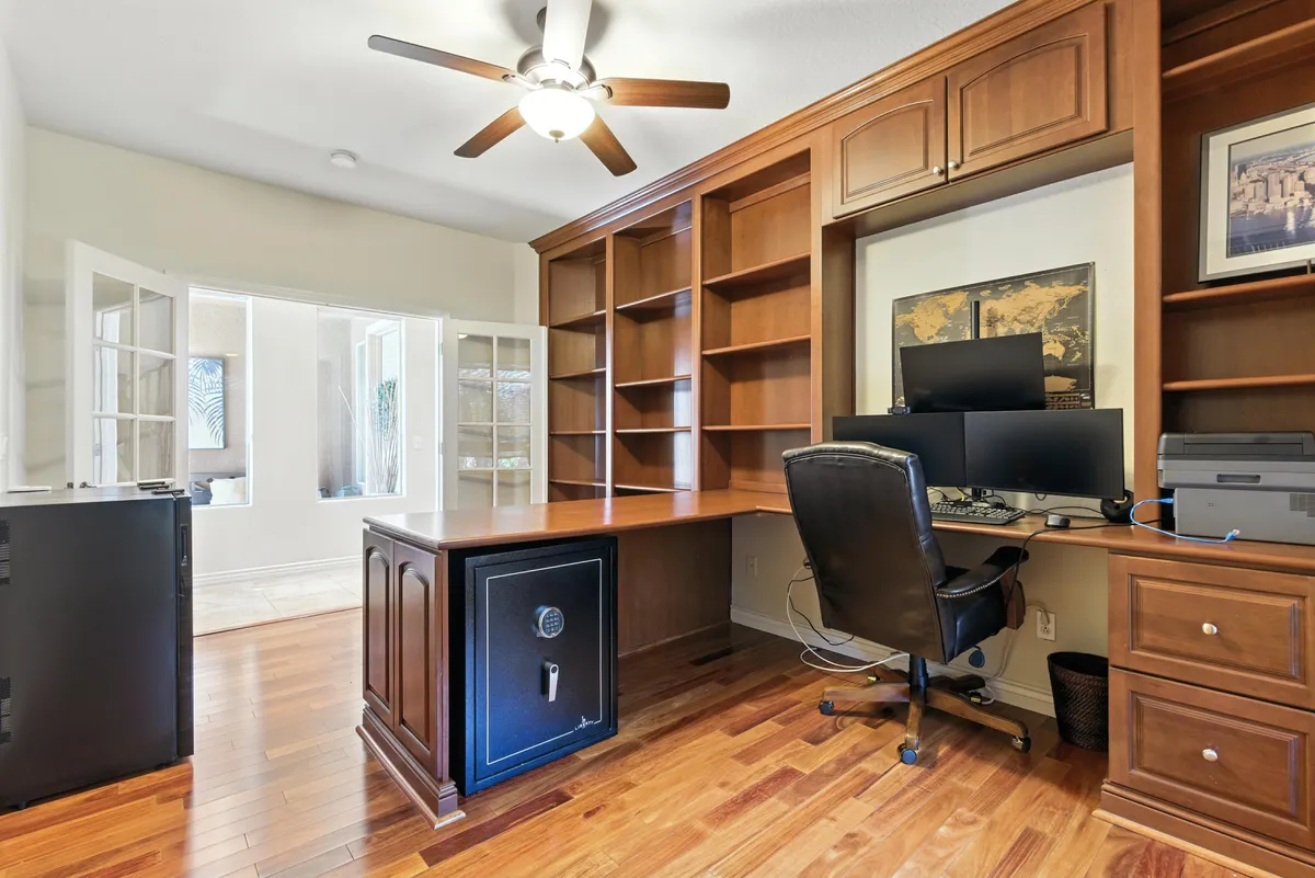 A bright, focused interior workspace featuring a wooden desk, built-in shelves, a computer setup, and ample natural light coming through french doors. The organized desk and inviting ambiance make this the ideal spot for video calls, collaborative sessions, or independent productivity.