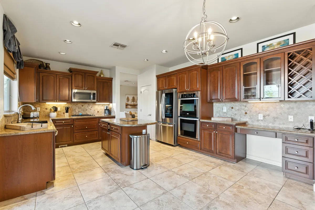 A spacious, stylish kitchen featuring a central island, gleaming stainless steel appliances (including a double oven), glass-front cabinets, and a welcoming chandelier overhead. The space is bathed in natural light, inviting creativity and togetherness.