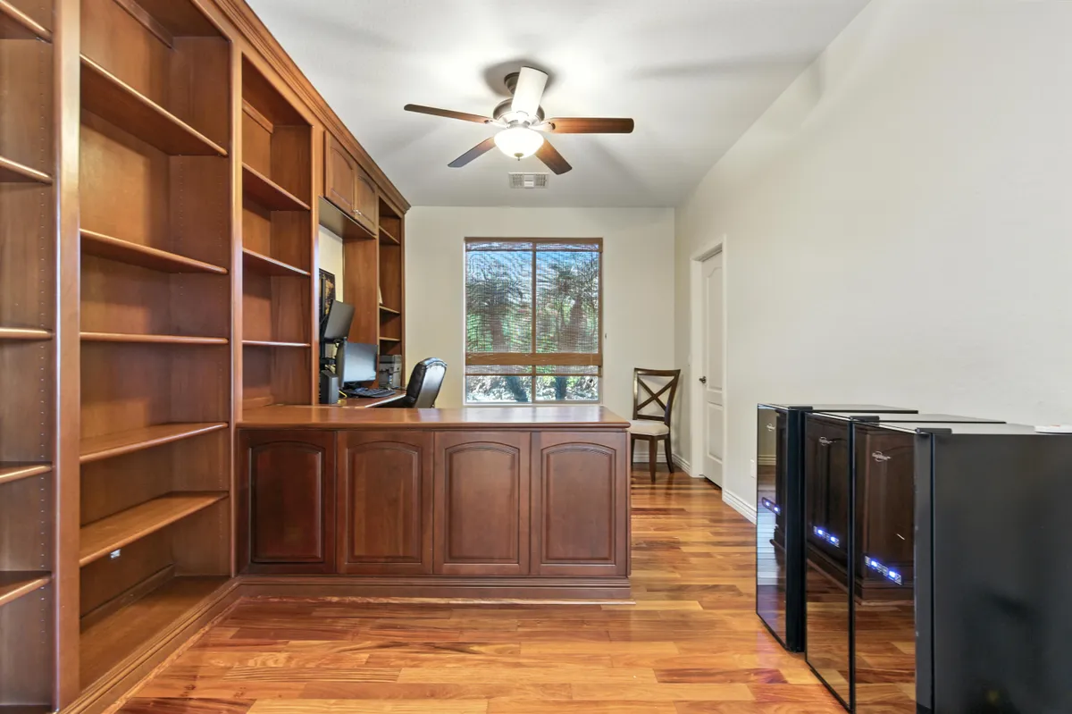 A dedicated computer room with a wooden desk, bookshelves, a computer setup, and a window providing serene views and ample natural light. Barstools offer flexible seating, and the space feels focused yet comfortable.