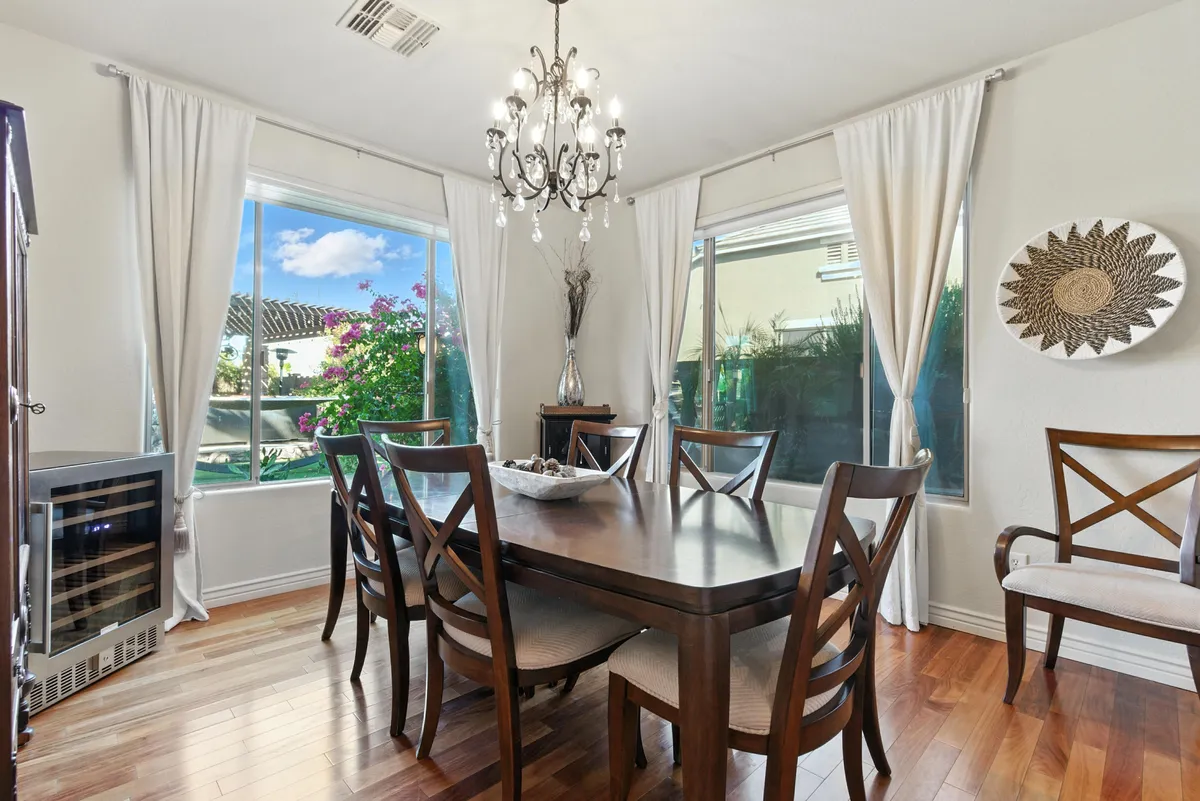An elegant dining room with a large table set for six, a beautiful chandelier above, and large windows letting in daylight. Decorative elements add warmth and sophistication, making this the ideal spot for group meals.