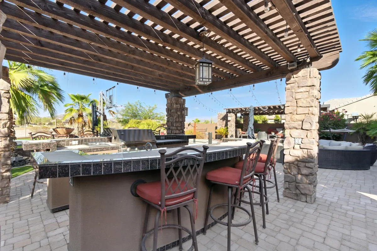 A stylish outdoor bar area beneath a pergola adorned with string lights, framed by stone pillars and palm trees in the background. There is bar seating perfect for enjoying drinks with friends as the sun sets.
