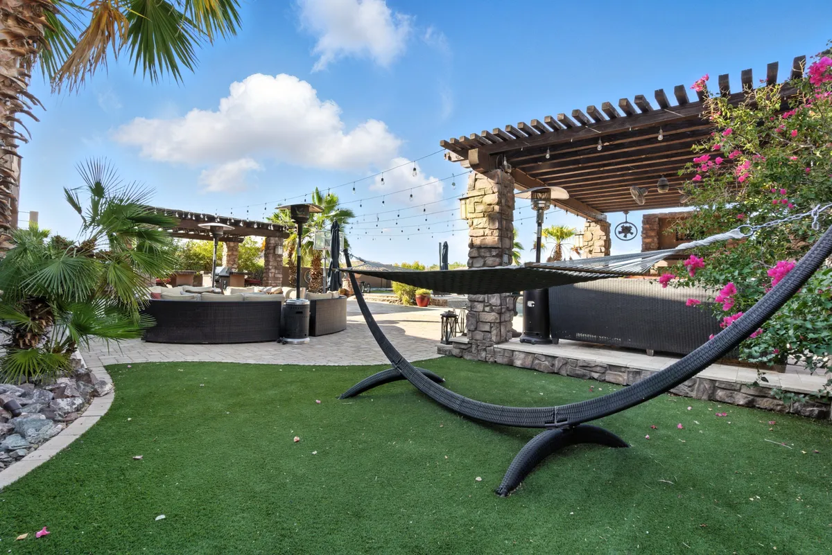 A tranquil backyard nook featuring a hammock strung between stone pillars, with lush greenery and string lights adding to the dreamy, relaxing setting. There’s also comfortable seating for lounging or socializing.