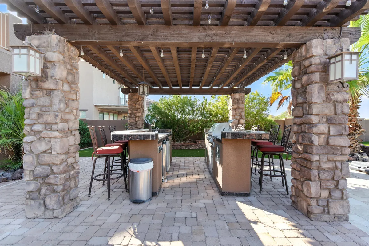 A spacious outdoor kitchen under a pergola, complete with bar stools, stone columns, and nearby entertaining spaces. The area is designed for both cooking and socializing, perfect for al fresco meals and celebration parties.