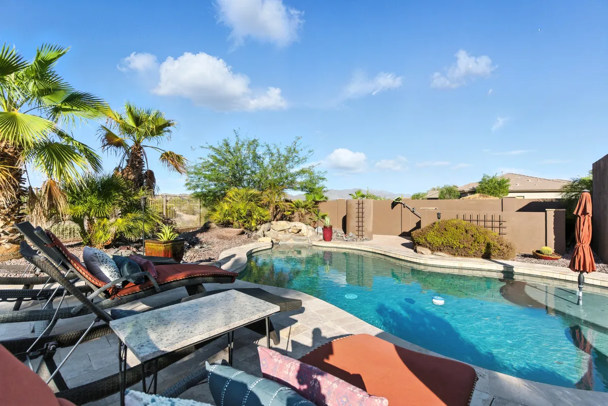 A tranquil outdoor swimming pool surrounded by lounge chairs, palm trees, and rock formations. The scene evokes relaxation and leisure, with the pool area bathed in natural light and offering views toward the landscaping.