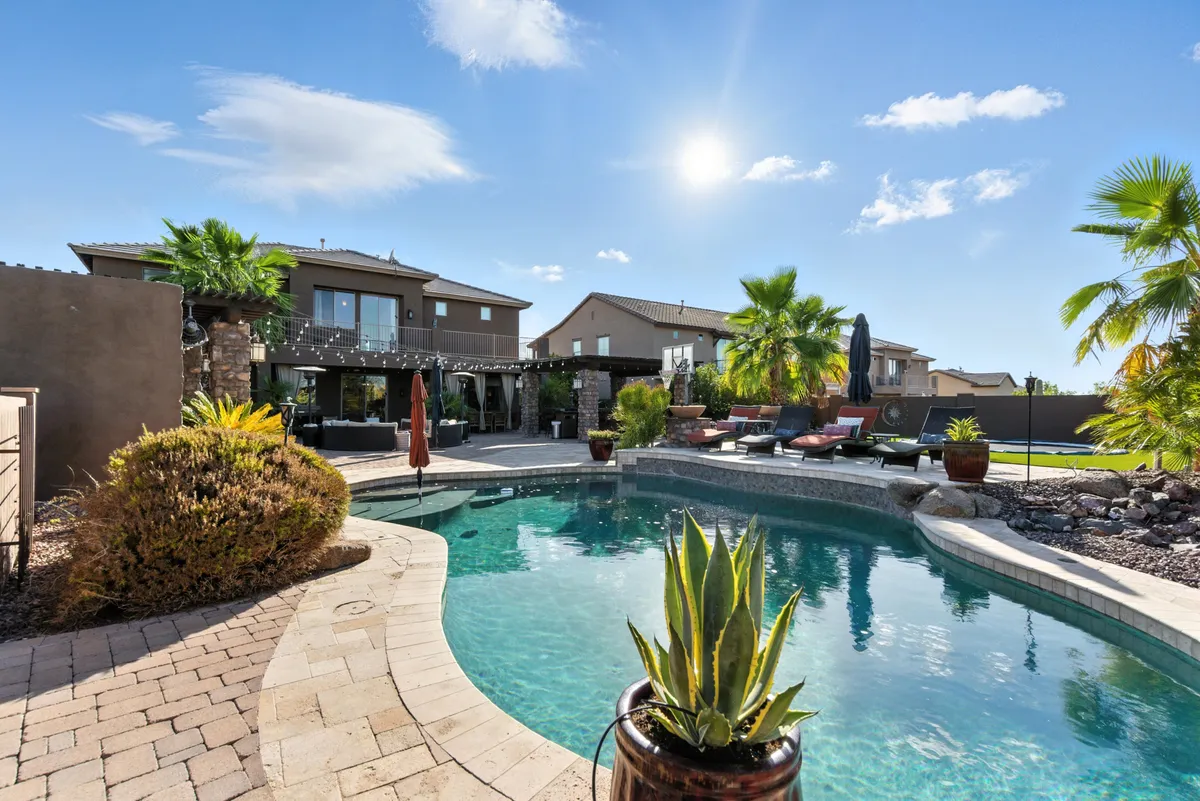 A vibrant outdoor scene featuring a resort-style swimming pool surrounded by lounge chairs, lush palm trees, and a welcoming outdoor seating area under a clear sunny sky. In the background, a fire pit hints at evening gatherings, while the spacious layout suggests opportunities for both relaxation and social connection.
