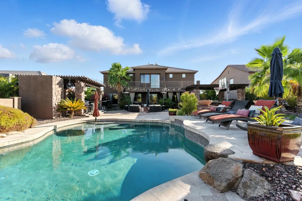 The image showcases a stunning backyard oasis at a single-family home in Goodyear, Arizona. The centerpiece is a sparkling swimming pool flanked by swaying palm trees, an inviting outdoor seating area perfect for gatherings, and lush, manicured landscaping. The scene exudes relaxation and luxury, with plenty of space for swimming, sunbathing, and entertaining under the desert sky.
